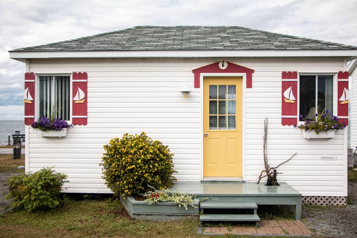 Une petite maison blanche avec une porte jaune, des volets rouges et un petit porche avec un banc.