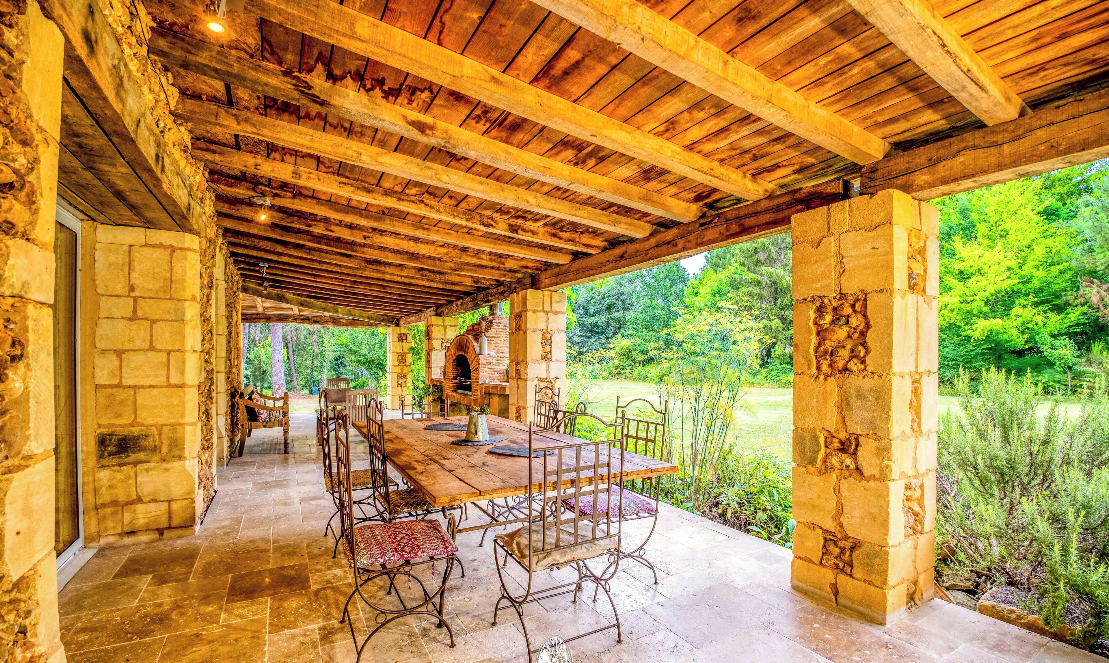Une terrasse extérieure couverte, dotée d’un mur en pierre, de poutres en bois et d’une table à manger avec des chaises.