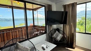 Et moderne værelse med stort TV, to sorte stole, et lille bord og udsigt til sø og bjerge.