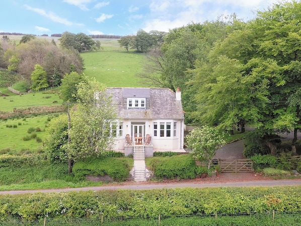 A white cottage with a stone staircase leading to a wooden door, surrounded by lush greenery and a fenced garden.