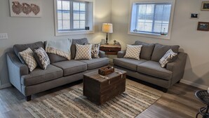 A living room with two grey sofas, a wooden coffee table, and a striped rug. There are two windows with blinds, a ceiling fan, and a lamp on a side table.