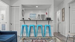 A modern kitchen with white cabinets, a stainless steel refrigerator, and a blue countertop. There are four blue bar stools and a glass coffee table in the center. - Tranquil Tortuga *Lake View* Heated Pool (Destin)