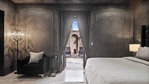 Een moderne slaapkamer met een groot bed, een donkere fauteuil, een kroonluchter en uitzicht op een buitenplaats met een bakstenen boog.