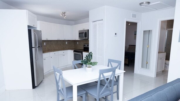 A modern kitchen with white cabinets, a stainless steel refrigerator, a microwave, and a dining area with a white table and blue chairs.