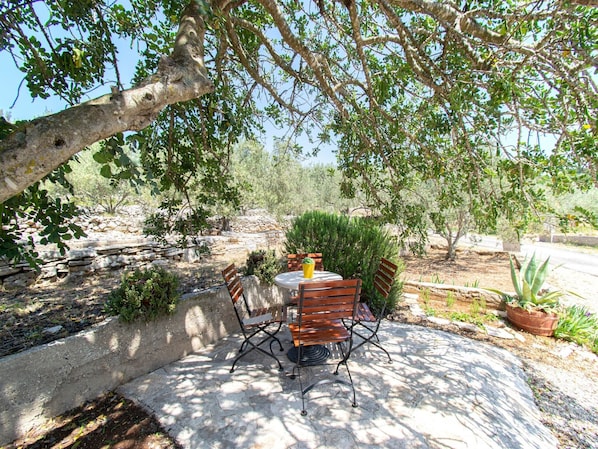 An outdoor seating area with a stone table and wooden chairs, surrounded by greenery and a stone wall.