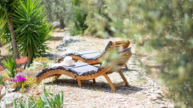 Twee houten loungestoelen met kussens, geplaatst op een grindpad omringd door groen en bloeiende planten.