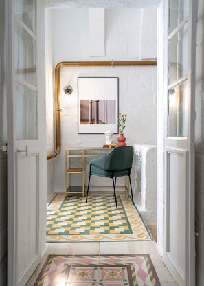 A narrow hallway with a checkered floor, a desk with a chair, a framed picture on the wall, and a vase with flowers.