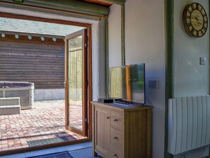 Une pièce avec un meuble en bois, un téléviseur posé dessus, une horloge murale et une porte-fenêtre coulissante donnant sur un espace extérieur avec une terrasse et un jacuzzi.