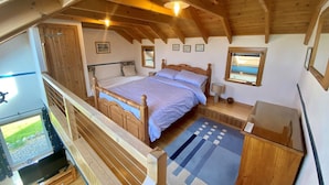 Une chambre avec un plafond en bois, un lit doté d’une tête de lit en bois, une table de chevet avec une lampe et une fenêtre donnant sur l’extérieur.