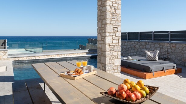A sunlit terrace with a stone pillar, a wooden table with breakfast, a lounge area with a mattress and pillows, and a view of the sea. - Southrock Villas - 6 Bed Magnificent Seaview Villa with Private Pool New Listing (Lachania)
