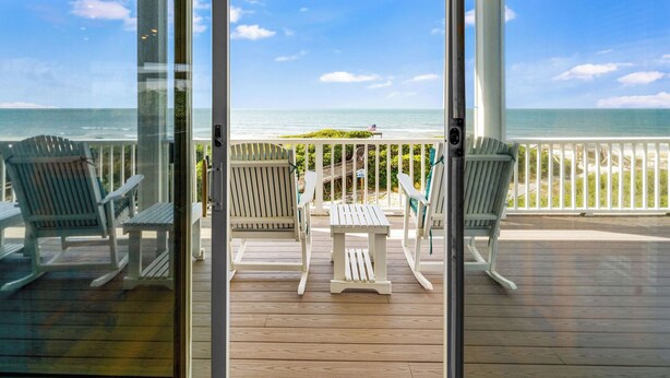 Une terrasse en bois avec des fauteuils Adirondack blancs et une petite table, donnant sur une plage et l’océan.
