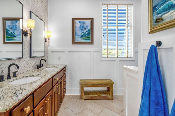 Une salle de bain avec un double lavabo, un meuble-lavabo en bois, deux appliques murales, une fenêtre avec stores et une serviette bleue accrochée au mur.