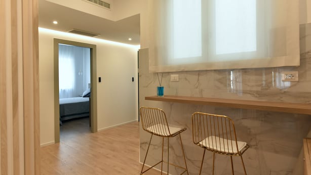 A modern room with a bed, a small sitting area with two chairs, a marble countertop, and a window with blinds. - Dimora al castello (Matera)