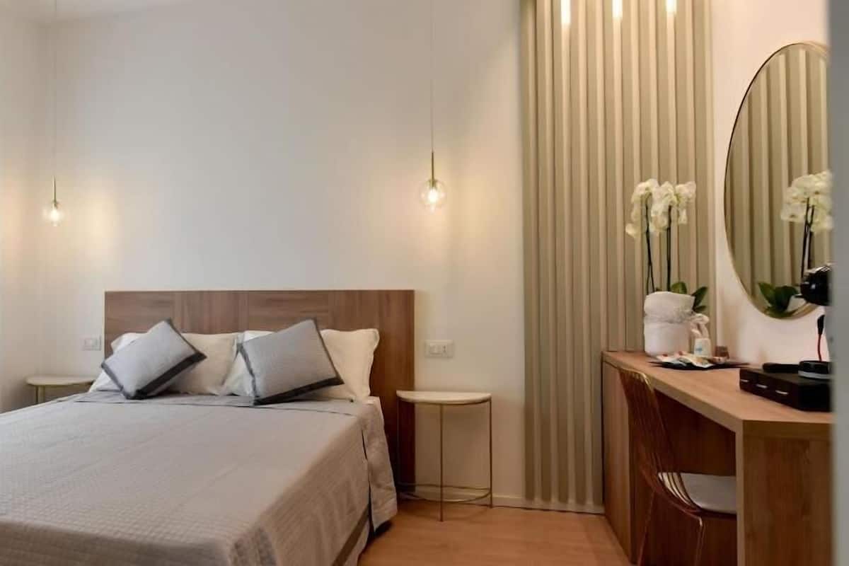 A hotel room with a bed, bedside tables, a desk, a mirror, and a radiator.