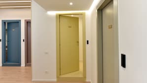 A modern hotel corridor with a yellow door, a blue door, and a closed elevator. - Dimora al castello (Matera)