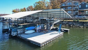 A dock with a covered seating area, a boat, and a house in the background. - BARE FEET RETREAT! Swim Dock, 5 Decks, Elevator, Gaming Room, Bagnell Dam Strip! (Lake Ozark)