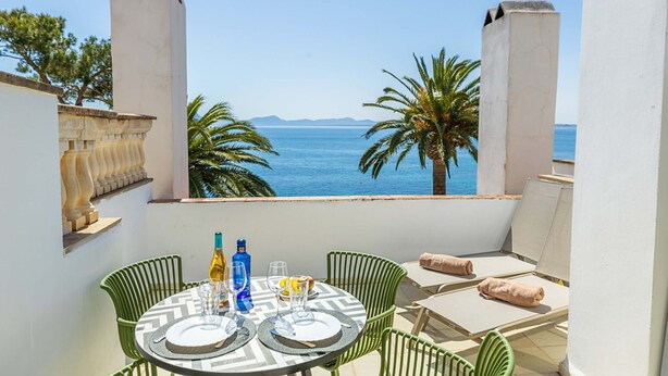A balcony with a table set for two, overlooking a coastal view with palm trees and the sea.