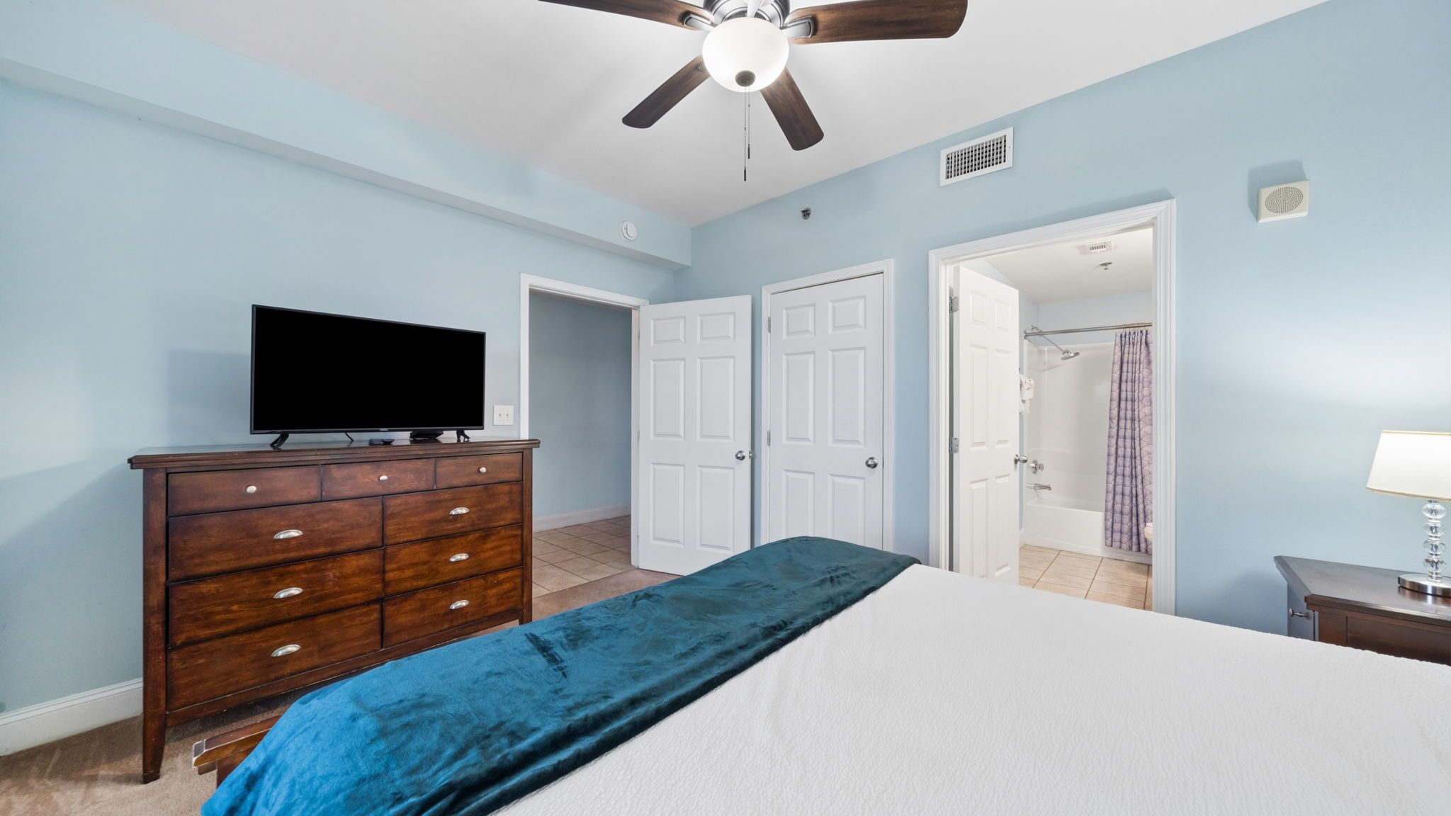 Une chambre à coucher comprenant un lit, une commode, un téléviseur, un ventilateur de plafond et une porte donnant sur une salle de bain.