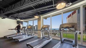 A gym with treadmills and weight machines, large windows with a city view, and a high ceiling with exposed beams. - Sand n Stars (Panama City Beach)