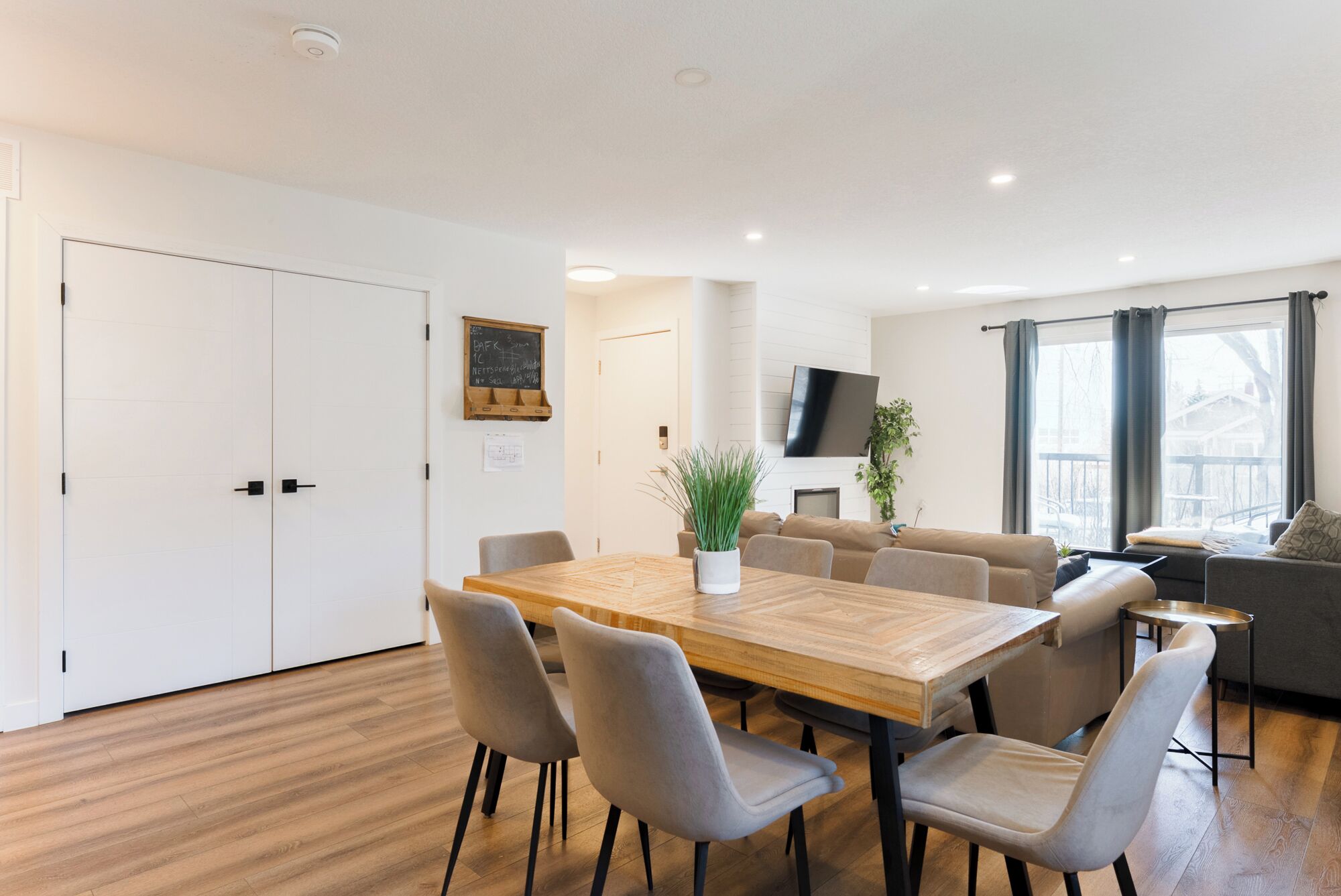 A modern dining area with a wooden table, grey upholstered chairs, a potted plant, and a flat-screen TV mounted on the wall.