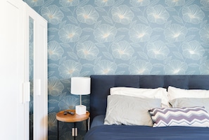 A bedroom with a floral wallpaper, a bed with multiple pillows, a bedside table with a lamp, and a remote control.