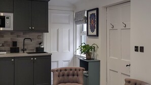 Ett modernt kök med mörka skåp, en mikrovågsugn och en diskho. I mitten av rummet står en stoppad stol, och en till stol står till höger. En krukväxt står på ett litet bord bredvid fönstret.