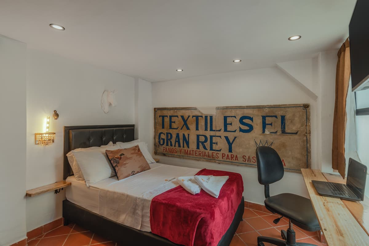 Une chambre à coucher avec un lit, un bureau avec un ordinateur portable, une chaise, une lampe fixée au mur et une enseigne sur laquelle on peut lire : « TEXTILES EL GRAN REY ».