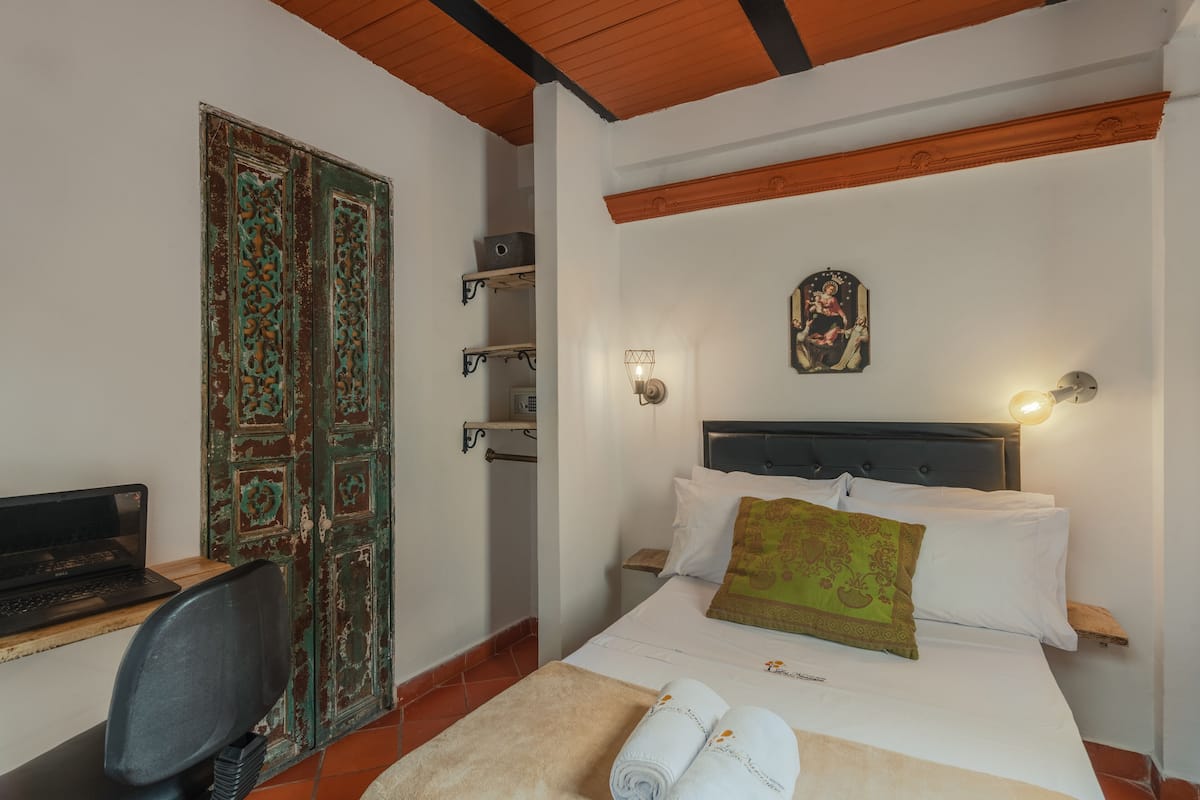 Une chambre à coucher avec un lit, un bureau avec un ordinateur portable, une chaise, une porte verte et un luminaire fixé au mur.