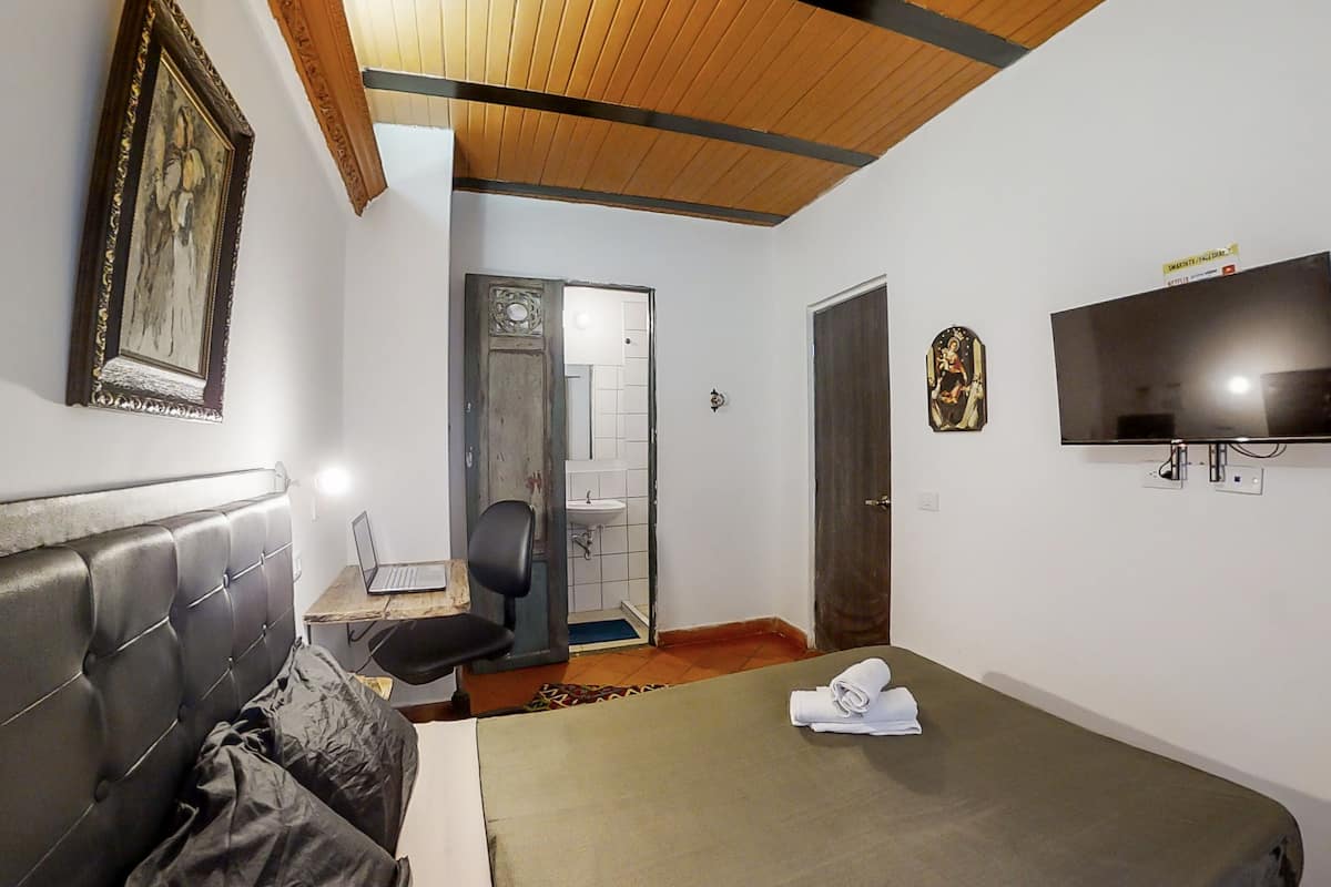 Une chambre d’hôtel avec un lit, un bureau avec un ordinateur portable, une télévision et une salle de bain visible à travers une porte ouverte.