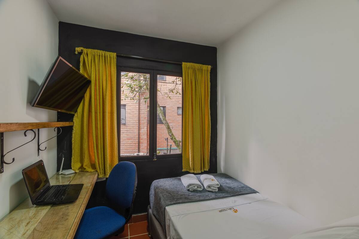 Une petite pièce, de taille modeste, comprenant un lit, un bureau avec un ordinateur portable, une chaise et une fenêtre avec des rideaux.