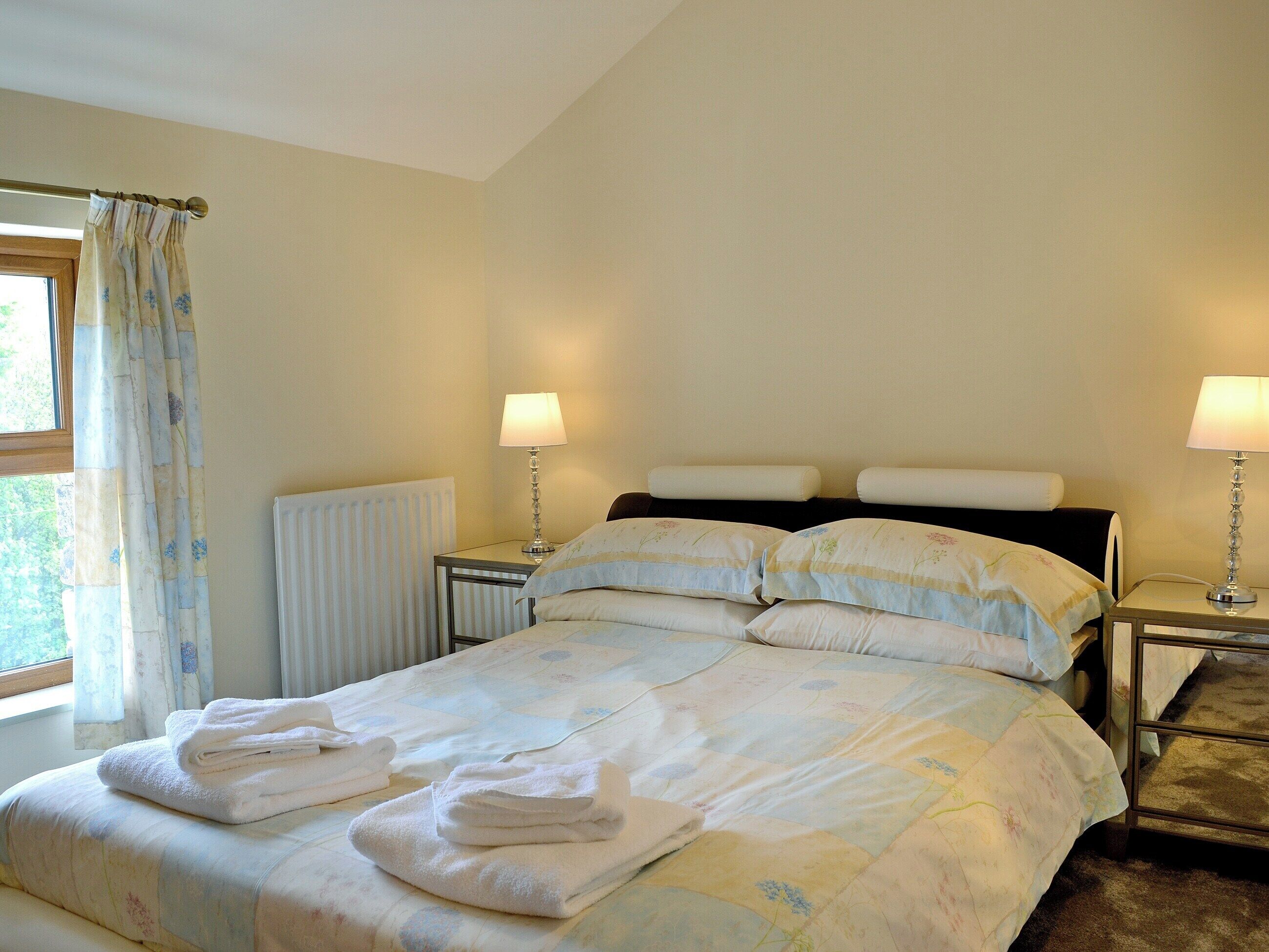 Una cama bien hecha con almohadas y toallas dobladas, una mesita de noche con una lámpara y una ventana con cortinas.