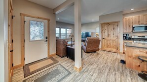 A spacious living area with a sofa, a coffee table, and a TV. There is a wooden door with a stained glass window, a kitchen with wooden cabinets, and a dining area with a table and chairs. - Midgarth Hus- Brand New PET FRIENDLY home minutes to Copper & Ski Cooper! (Leadville)