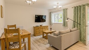 A living room with a wooden dining table, chairs, a sofa, a TV, and a window with curtains. - 1 bedroom accommodation in Ledbury (Ledbury)