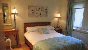 A neatly made bed with a green quilt, two bedside lamps, a wooden nightstand, a mirror, and a window with curtains.