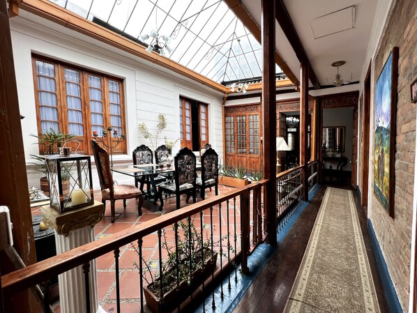 A spacious indoor area with a high ceiling and a glass roof, featuring a dining table with chairs, a wooden railing, and potted plants. - Hotel Galería Casa de La Luz   (Bogotá)