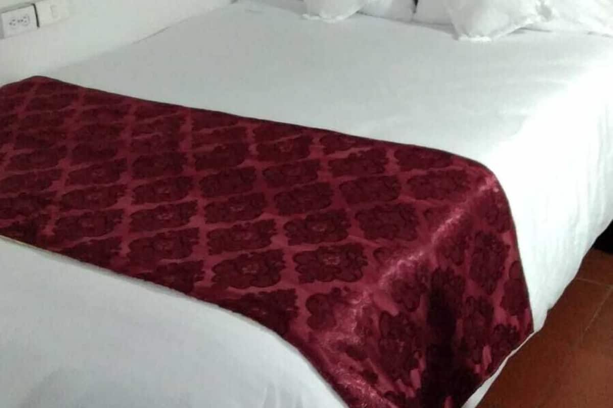 A hotel room with a bed, white bedding, a red patterned throw, green curtains, and a painting on the wall.