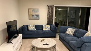 Ein Wohnzimmer mit Fernseher auf einem weißen Schrank, einem runden Couchtisch und zwei blauen Sofas mit gemusterten Kissen.