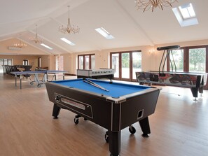A pool table with a blue felt surface, a foosball table, and a ping pong table in a spacious room with wooden flooring and skylights.