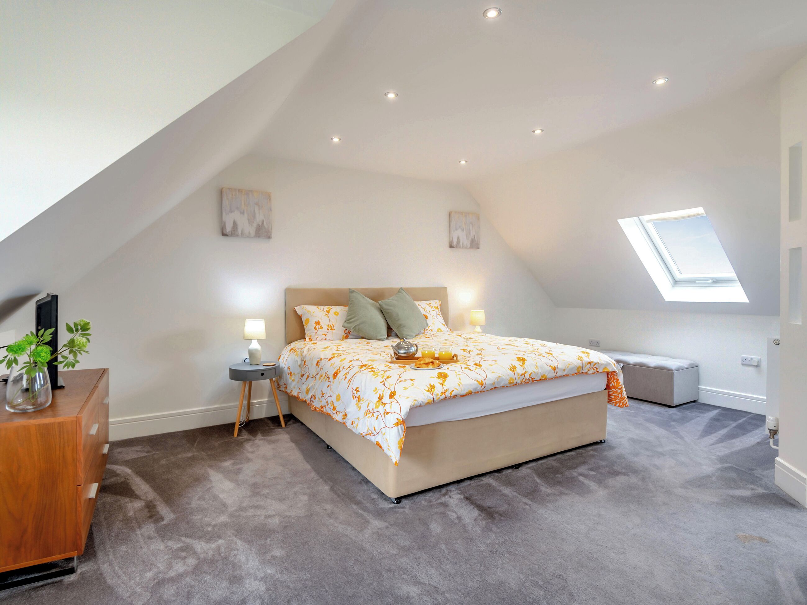A bedroom with a sloped ceiling, a bed with a floral bedspread, a bedside table with a lamp, a small plant, and a window.
