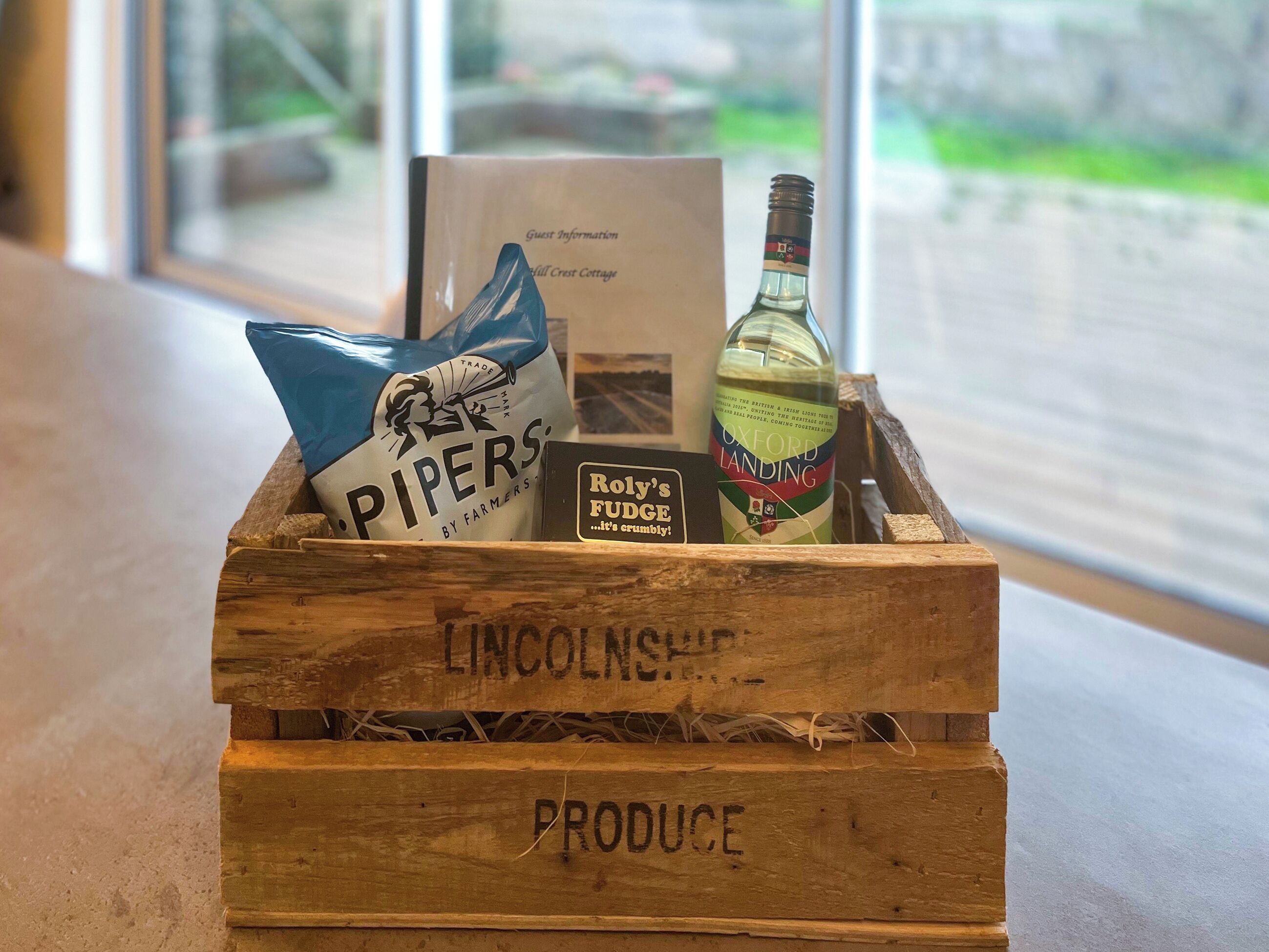 Una caja de madera con la etiqueta 'LINCOLNSHIRE PRODUCE' que contiene una bolsa de 'PIPERS BY FARMERS', una botella de 'OXFORD LANDING' y un cartel de 'Roly's FUDGE'.
