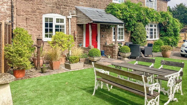 A stone building with a red door, a small porch, and a well-maintained garden with a bench. - A delightful cottage offering comfortable accommodation for up to four guests. (Much Birch, near Hereford)