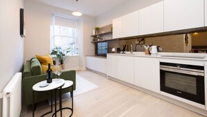 A modern kitchen with white cabinets, a built-in oven, and a dining area with a green sofa and a small table. - Trendy Studio on Rose South (Edinburgh)