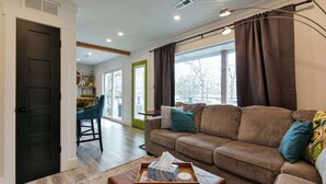 A living room with a sofa, a coffee table with a tissue box, a dining area with a chair, and large windows with curtains.