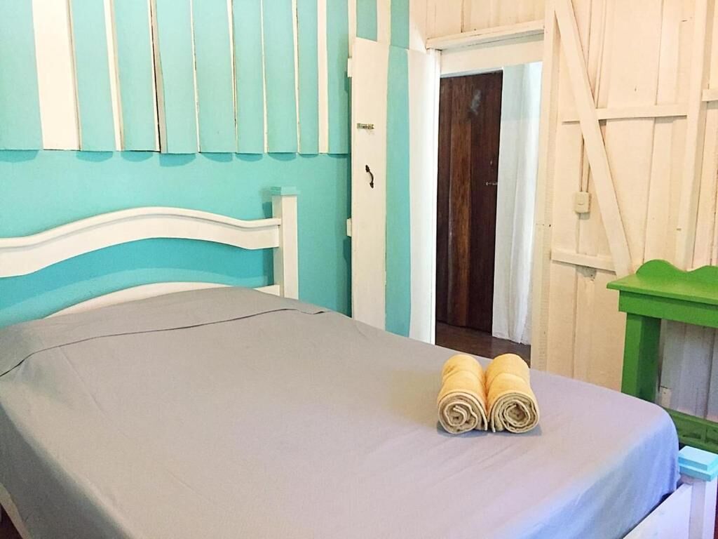 Una cama con sábana gris y dos toallas enrolladas, cabecera blanca y una mesita verde a la derecha.