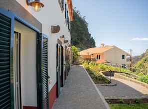Une rangée de portes fermées, dotées de volets, un chemin pavé et un espace jardin agrémenté de plantes et d’un petit élément décoratif aquatique.