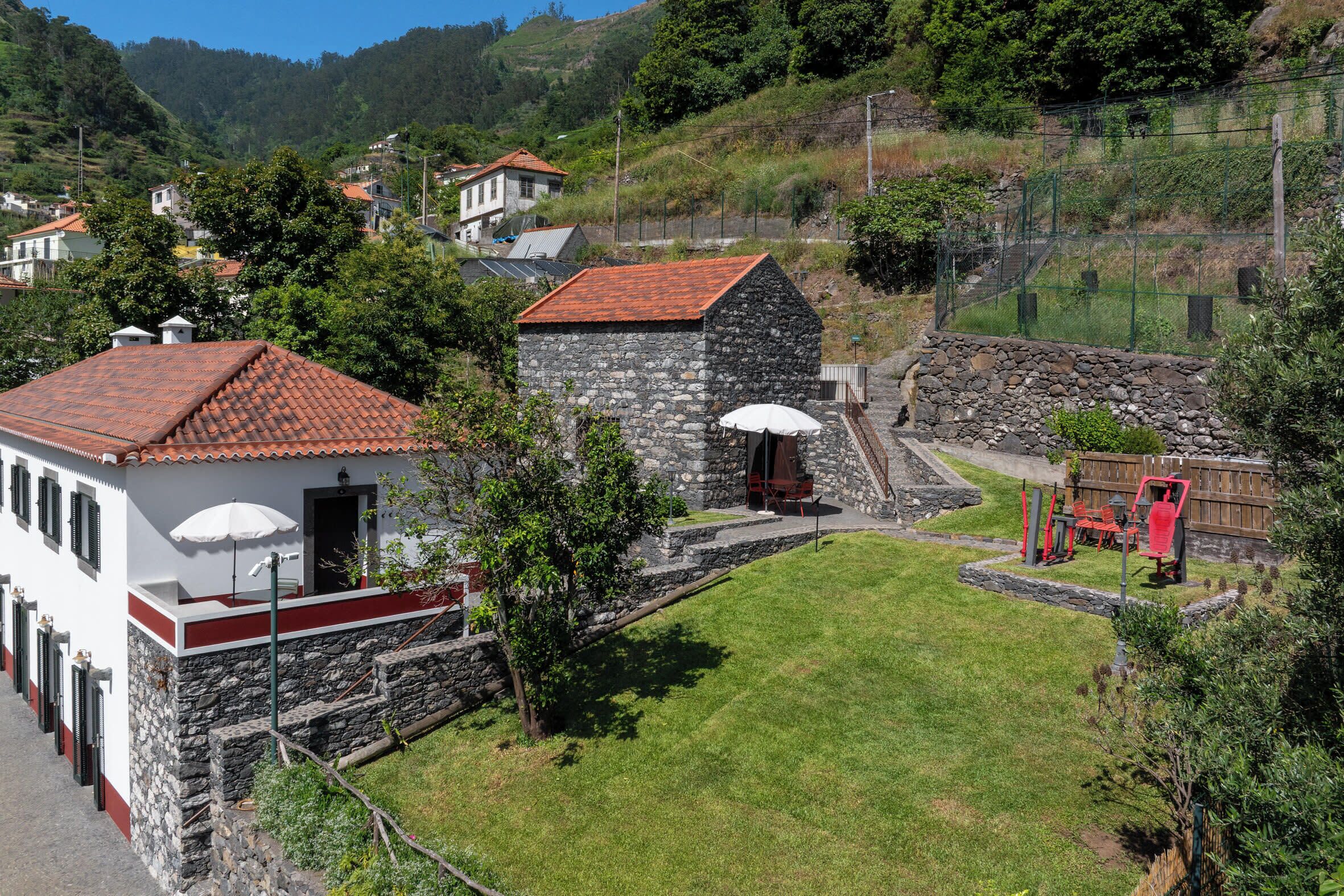 Un bâtiment en pierre avec un toit recouvert de tuiles rouges, un bâtiment blanc avec un balcon rouge et un espace verdoyant aménagé avec du mobilier d’extérieur.