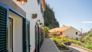 Una hilera de puertas cerradas con contraventanas, un camino empedrado y un jardín con plantas y una pequeña fuente.