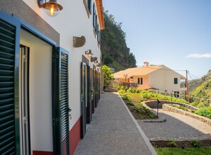 A row of closed doors with shutters, a cobblestone pathway, and a garden area with plants and a small water feature.