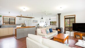 A spacious living area with a sofa, coffee table, and a TV. There is a kitchen area with a microwave and refrigerator. A fireplace is visible in the corner. - Callala Calm - Pet Friendly - 5 Mins Walk to Beach (Callala Bay)