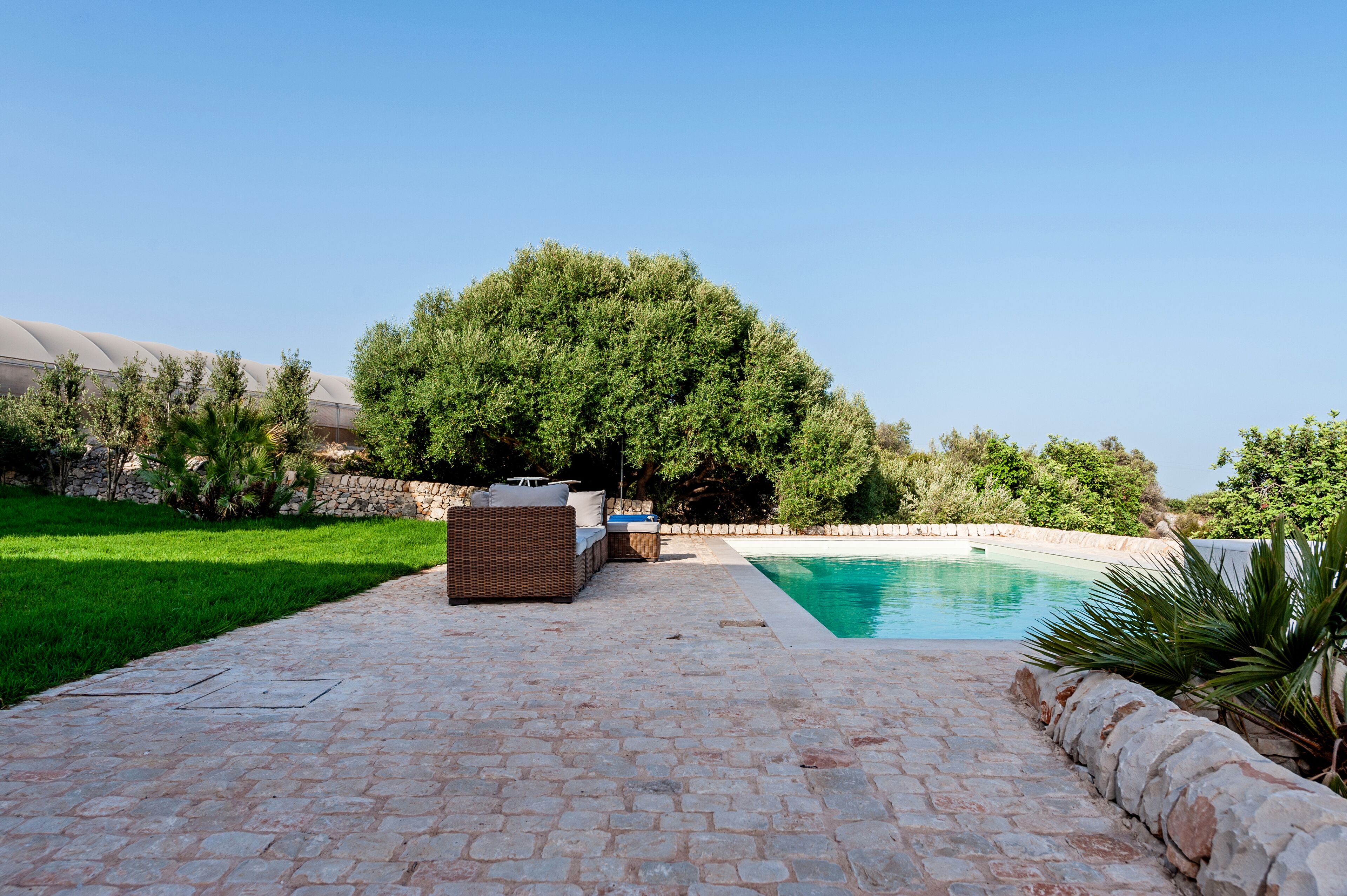 A pool area with a stone patio, a wicker sofa, and a large tree.